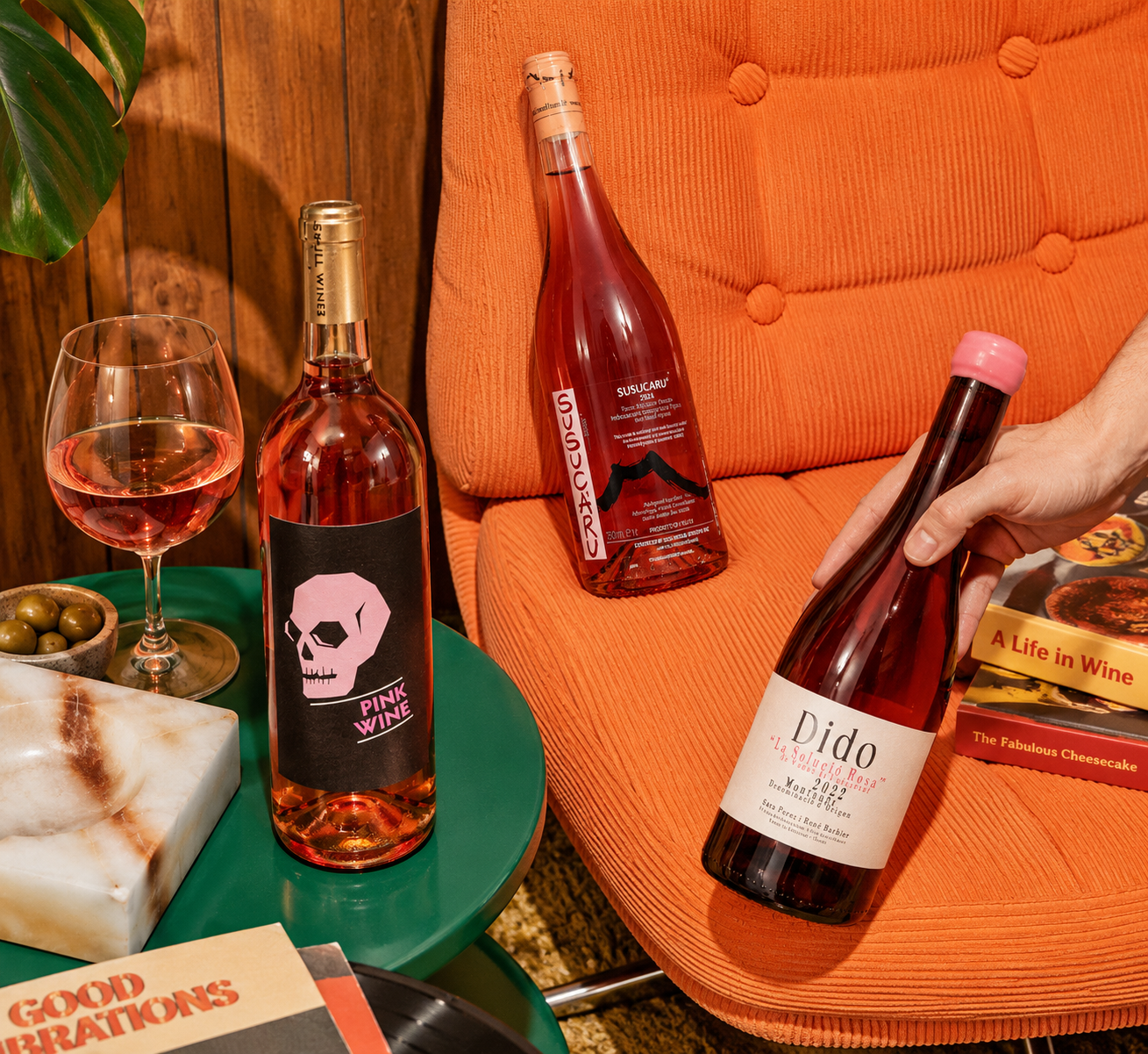 Two rosé wine bottles and a filled wine glass arranged on a green side table next to an orange upholstered chair. A hand holds one bottle labeled 'Dido Rosé' while another bottle labeled 'Pink Wine' with a pink skull graphic stands on the table