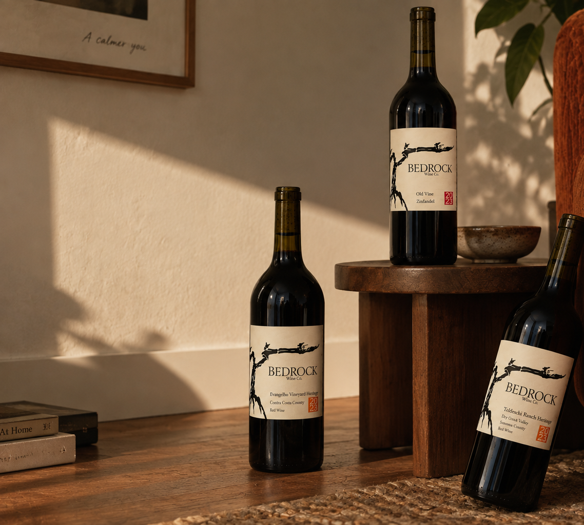 Three bottles of BEDROCK wine displayed on a wooden table and stool in a warm, sunlit room setting.