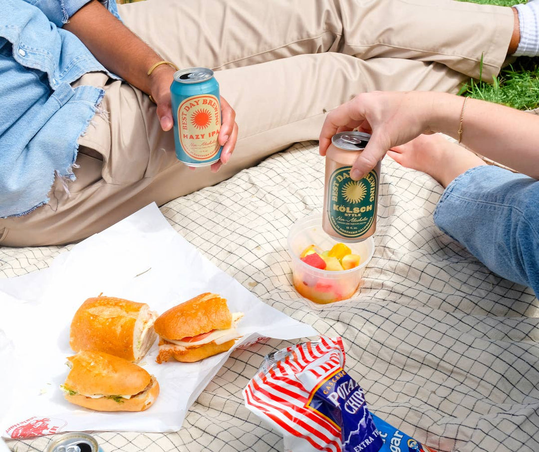 Two people enjoying a picnic with Best Day Brewing non-alcoholic beer, sandwiches, and a fruit cup on a blanket outdoors.