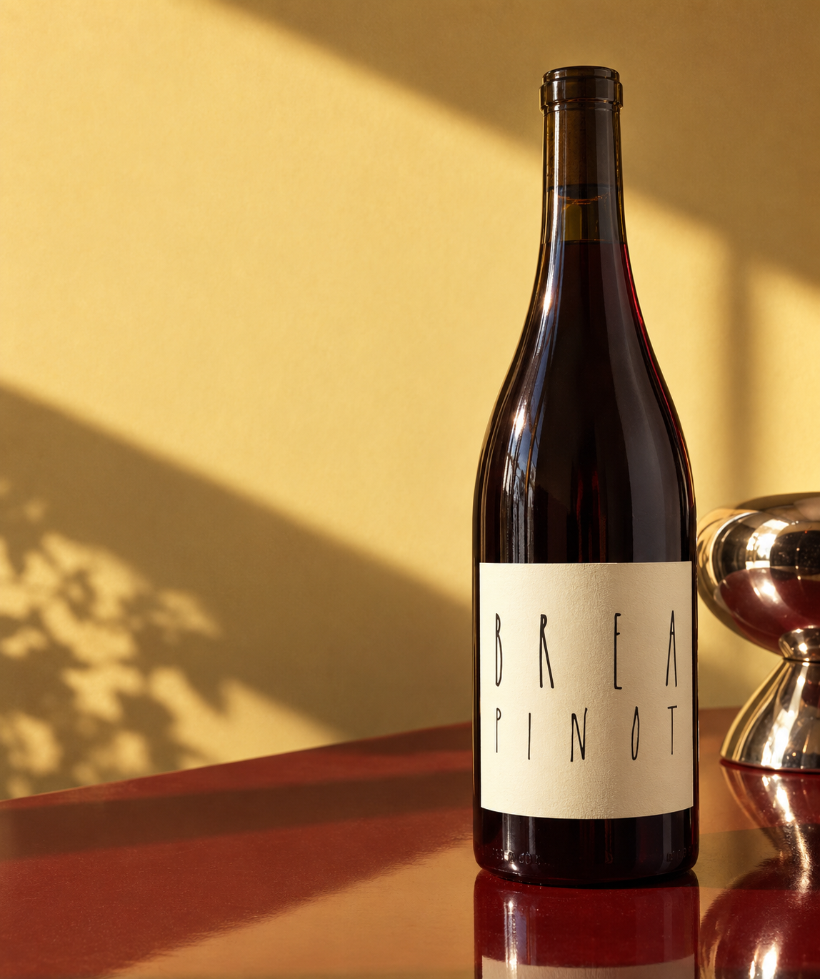 A dark glass bottle of Brea Pinot wine with a cream label, standing on a reflective red surface against a warm yellow wall with dramatic shadows.