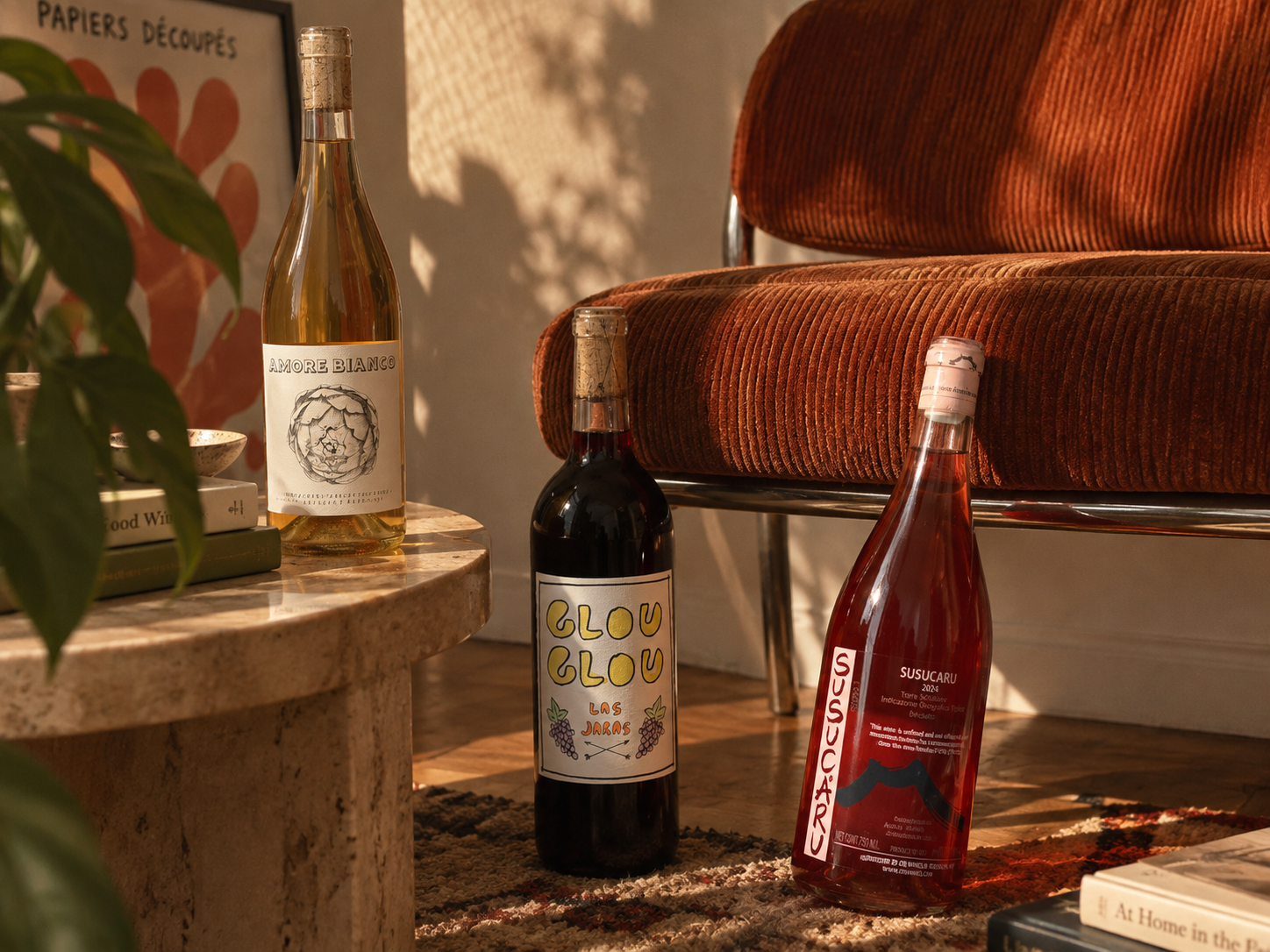 Three wine bottles from the brand Broc Cellars, Frank Cornelissen, and Las Jaras are displayed in a cozy, sunlit room setting. One bottle of white wine sits on a stone side table next to a plant, while two other bottles stand on the floor in front of a rust-colored corduroy couch.