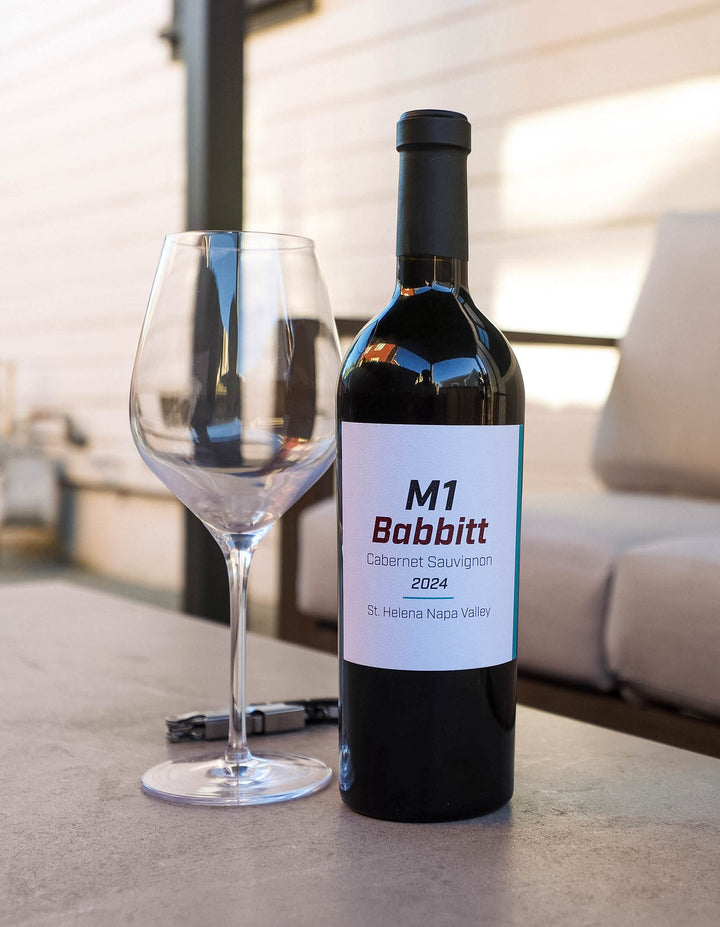 A bottle of M1 Babbitt Cabernet Sauvignon 2024 wine next to an empty wine glass on a stone patio table, with a blurred outdoor patio background.