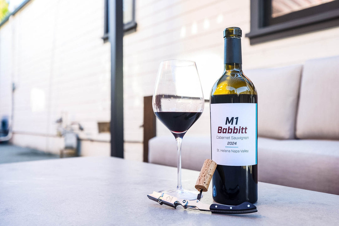 A bottle of M1 Babbitt Cabernet Sauvignon 2024 wine, a filled wine glass, a corkscrew, and a cork on a light gray table in an outdoor patio setting.